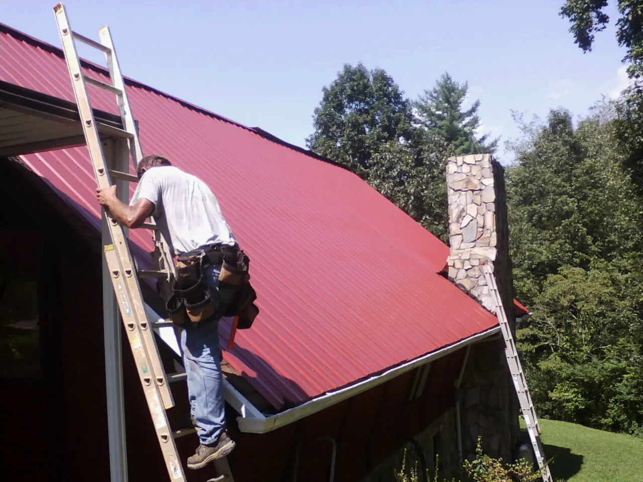 Roofer on a ladder beside a red metal roof and stone chimney — Soffit & Fascia Repair by Roofing Brothers in Naches